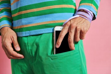 Person wearing green pants and a colorful striped long sleeve shirt putting a smartphone into their pocket against a pink background