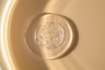 Clear transparent gel droplet with small bubbles inside reflecting soft warm light on beige surface