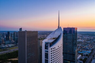 Modern cityscape at sunset featuring three prominent skyscrapers with one uniquely shaped building having a tall spire, urban landscape, and distant horizon under a clear sky