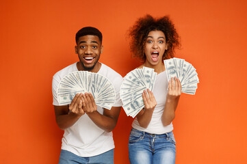 Big win! Shocked and happy black guy and girl holding lots of money and shouting, orange background