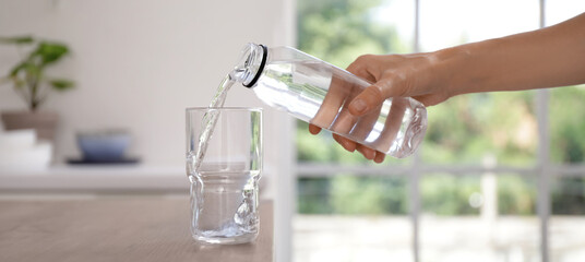 Woman pouring water from bottle into glass on table in kitchen, closeup