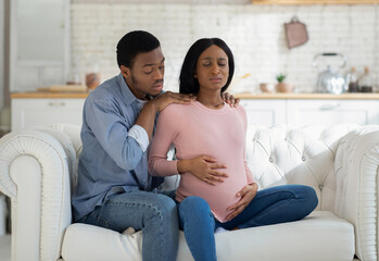 Worried black husband supporting pregnant wife with prenatal contractions at home. Beautiful African American woman suffering from pain, giving birth to baby with help of her partner