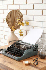 Vintage typewriter with hourglass, eyeglasses and vase on brown wooden table near brick wall