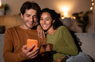 Cheerful Couple Using Cellphone With New Application, Wife Pointing Finger At Phone Browsing Internet Sitting On Couch At Home. Spouses Recommending Great App. Gadgets Concept