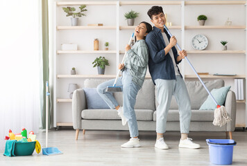 Happy asian couple singing songs and dancing while cleaning living room, using broom and mop as...
