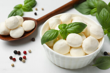 Tasty mozzarella cheese balls in bowl, spoon, basil and peppercorns on white marble table, closeup