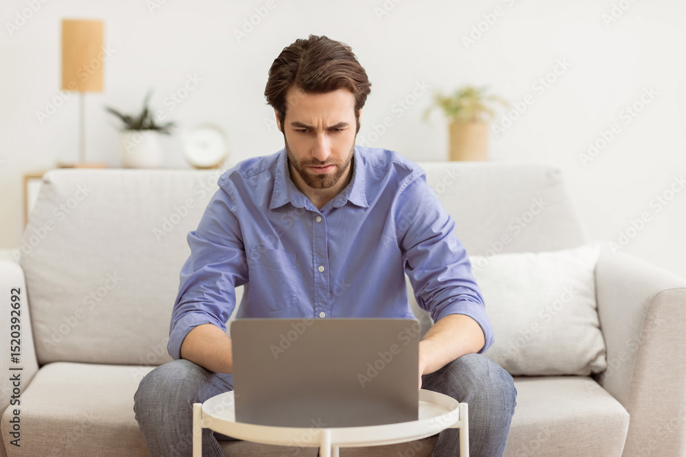 Poster man frowning using laptop computer having problem browsing internet sitting on sofa at home. unemplo