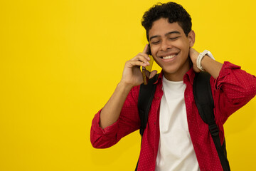 Young brown skinned Latino student wearing red formal wear and a black bag talking happily on his cell phone on a yellow background with copy space