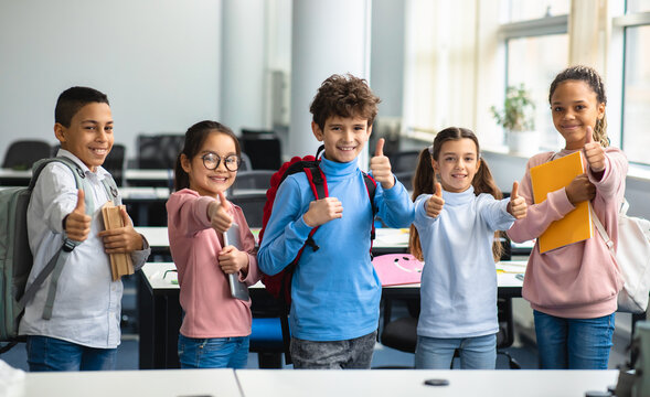Education Concept. Diverse group of happy smiling international classmates standing in line with notebooks and backpacks in classroom and showing thumbs up sign gesture, enjoying studying at school