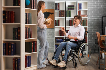 Male graduate in wheelchair talking to his friend with books at library
