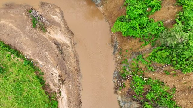Overhead shot reveals a muddy riverbank eroded by a recent landslide, emphasizing the role of deforestation and heavy rain in destabilizing slopes and increasing flood risks.
