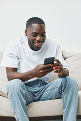Smiling young man using smartphone at home, wearing casual white t shirt and jeans, creating a...
