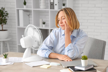 Beautiful mature sleepy businesswoman with electric fan suffering from heat in office