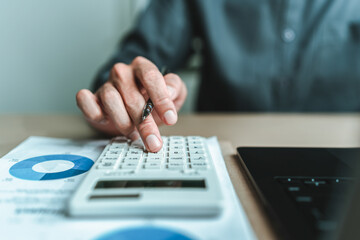 An accountant is seated at a desk in the office, utilizing a calculator to determine balances, profits, currency conversions, and costs. This involves various accounting and financial concepts.