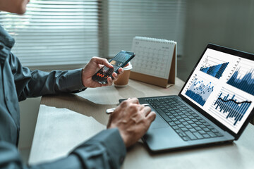 Businessman uses mobile phone to check stocks, news, email, market information while working with laptop screen graph, chart in Office.