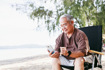 Happy elderly man using smartphone while sitting on chair at summer beach camping activity with family outdoors nature background