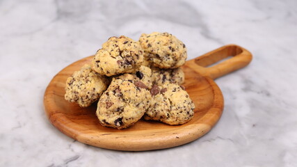A close-up image of a pile of rustic-style chocolate chip cookies placed on a round wooden serving plate with a handle. The cookies have a homemade, chunky appearance with visible chocolate chunks and