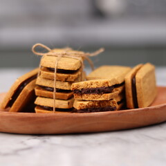 A close-up of handmade chocolate sandwich cookies tied with rustic twine, neatly stacked on a wooden plate. The golden brown biscuits with rich chocolate filling evoke a warm, homemade feel. Styled in