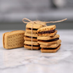 A close-up of handmade chocolate sandwich cookies tied with rustic twine, neatly stacked on a wooden plate. The golden brown biscuits with rich chocolate filling evoke a warm, homemade feel. Styled in