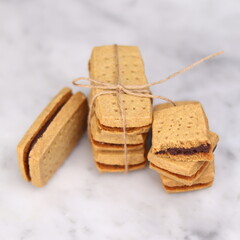 A close-up of handmade chocolate sandwich cookies tied with rustic twine, neatly stacked on a wooden plate. The golden brown biscuits with rich chocolate filling evoke a warm, homemade feel. Styled in