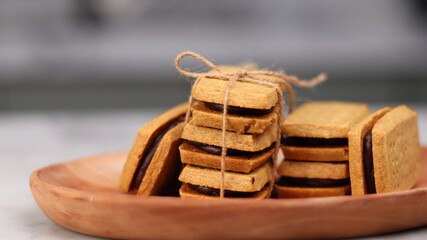 A close-up of handmade chocolate sandwich cookies tied with rustic twine, neatly stacked on a wooden plate. The golden brown biscuits with rich chocolate filling evoke a warm, homemade feel. Styled in