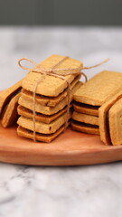 A close-up of handmade chocolate sandwich cookies tied with rustic twine, neatly stacked on a wooden plate. The golden brown biscuits with rich chocolate filling evoke a warm, homemade feel. Styled in