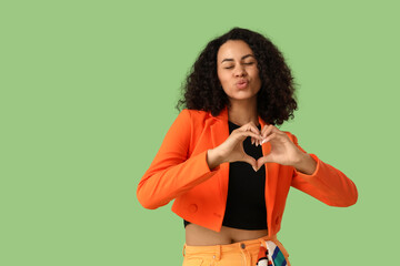 Stylish young African-American woman in orange blazer making heart gesture on green background