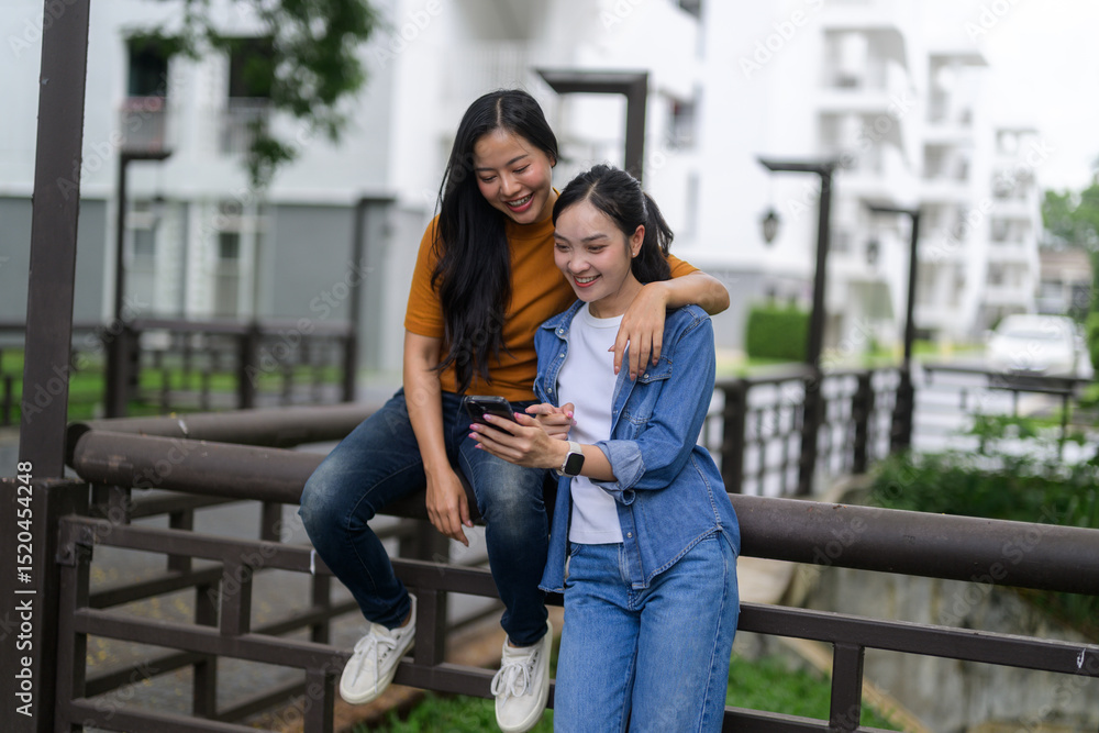 Poster two young women using mobile phone and smiling while sitting on a railing
