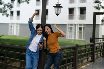 Two happy friends celebrating success while looking at good news on smartphone near bridge in the city
