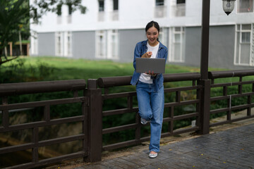 Young asian student pointing at laptop standing on bridge in university campus