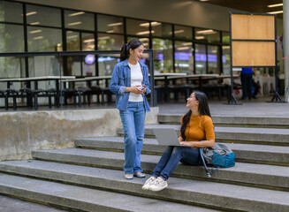 Two university students are discussing and working on a project on campus stairs, using a laptop...