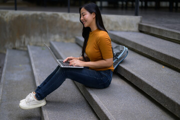 Smiling young Asian woman using laptop sitting on campus stairs, enjoying online learning and studying outdoors