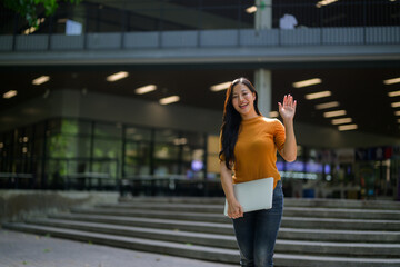 Happy asian student waving while holding laptop in university campus