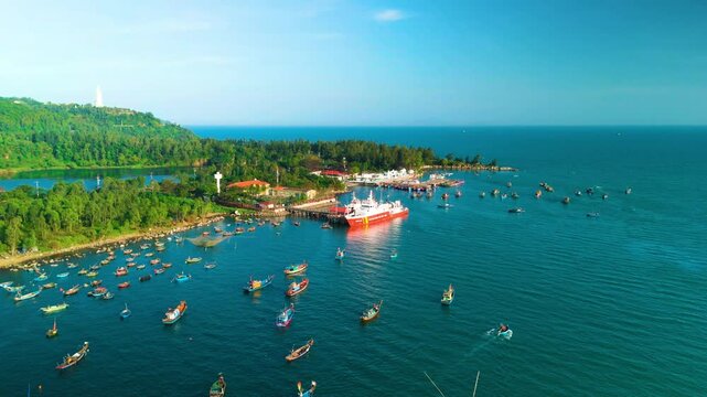 Colorful fishing boats navigating coastal waters near vung tau, vietnam, with large red white cargo vessel anchored alongside pier under bright blue sky