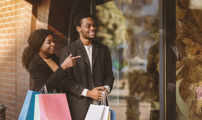 Cool sale in brand store, i want it. Interested african american guy and lady with colored paper bags walking to street, female shows finger at shop window at season sale in boutique, copy space