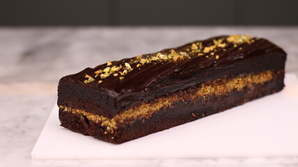 Rich chocolate layer cake topped with glossy dark chocolate ganache and sprinkled with chopped pistachios, presented on a white board over a marble surface. Elegant homemade dessert perfect for partie
