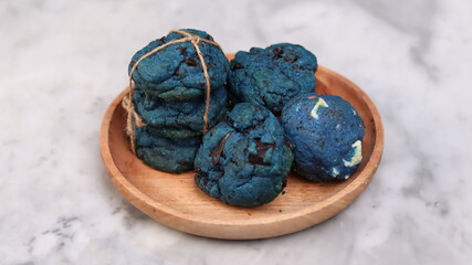 Vibrant blue chocolate chip cookies stacked and bundled with twine, served on a round wooden plate over a marble background. Eye-catching artisanal cookies perfect for bakery showcases, creative desse