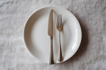A minimalist table setting featuring a white plate with a silver knife and fork placed on a light-colored cloth background