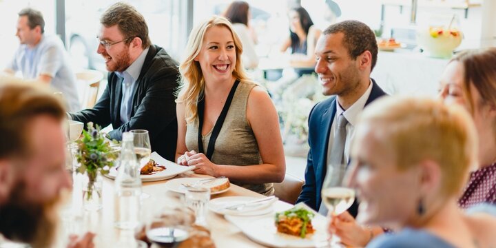 A diverse group of professionals, including a smiling blonde woman and a suited man, enjoy a lively lunch together. The atmosphere is warm and inviting, perfect for networking and socializing.