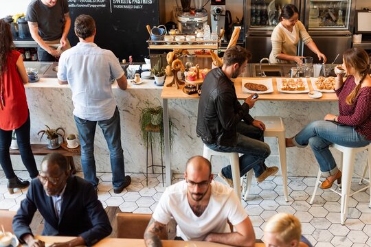 A busy cafe with diverse people enjoying coffee and pastries. Customers sit and chat, while baristas prepare drinks. A lively, social cafe atmosphere. Diverse people at a coffee shop. - Powered by Adobe