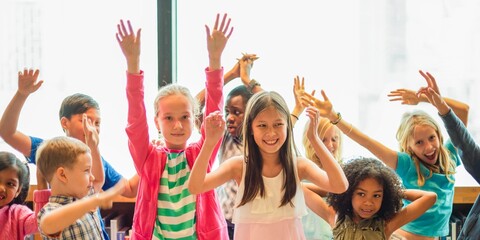 Group of diverse children joyfully raising hands in a classroom. Kids smiling, hands up, diverse group, classroom setting, joyful children, learning environment. Cute little diverse kids, arms raised.