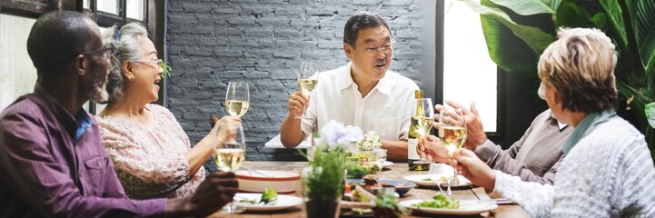 Group of diverse senior people enjoying a meal together. Diverse old people raising glasses in a toast. Smiling diverse elderly people celebrating around a table with food and drinks.