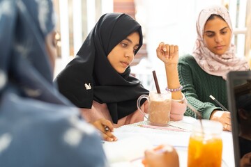 Three muslim girls in hijabs study together, focused on notes and drinks. Diverse group of women...