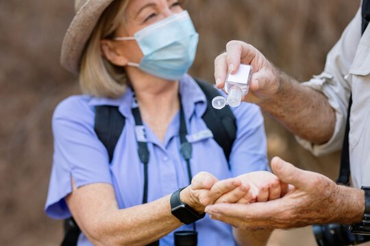 Senior woman wearing a mask receives hand sanitizer from a man. The woman, in a mask, enjoying outdoors. Senior woman using hand sanitizer. Senior couple enjoying outdoors adventure - Powered by Adobe