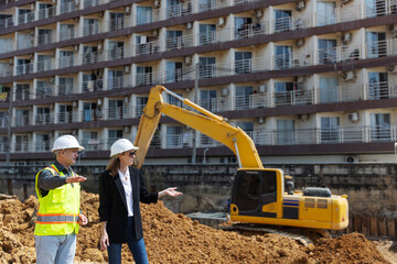 Two construction professionals in safety gear discuss and point toward excavation activity with a...