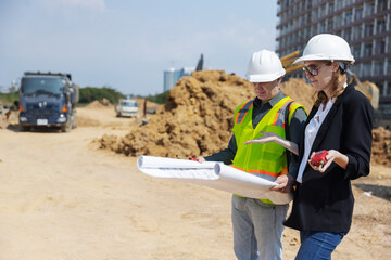 Two construction professionals review blueprints on a busy urban site surrounded by dirt piles, excavators, trucks, and large drainage pipes.