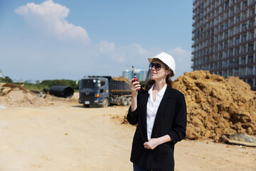A female construction manager wearing a hard hat and sunglasses communicates via walkie-talkie at an active job site with dump trucks, drainage pipes, and dirt piles in the background.
