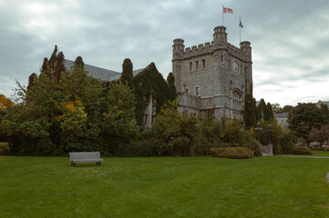 Ivy-covered historic stone building with a clock tower and Canadian flag. Lush greenery and cloudy sky create a moody and timeless campus or heritage site atmosphere.