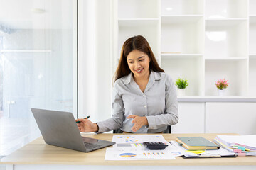 A woman is working on a desk in front of her with a pile of documents, a laptop, a calculator, a mobile phone and a notebook. This image shows her working on a project or analyzing data.
