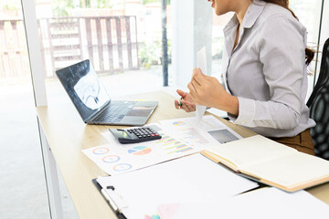 A woman is working on a desk in front of her with a pile of documents, a laptop, a calculator, a mobile phone and a notebook. This image shows her working on a project or analyzing data.
